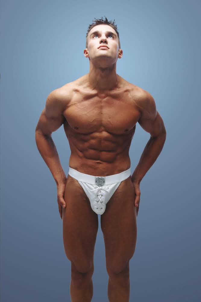 A studio fitness photograph of a muscular male model posing in white designer athletic underwear against a solid blue-grey background.